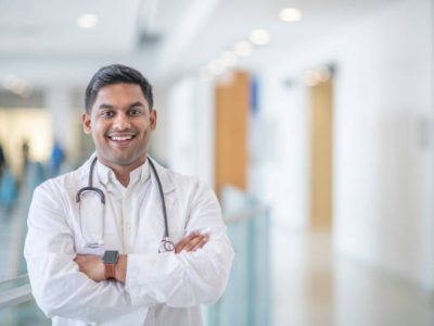 istockphoto-1342708859-612×612 A male doctor of Indian decent, smiles in this medical portrait. His arms are crossed as he stands in a hallway..